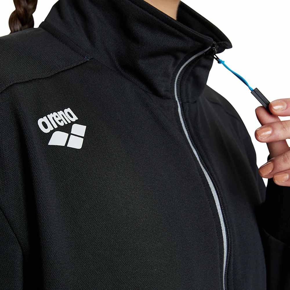 Close-up of a black Arena athletic jacket with a white logo on the chest, featuring a high collar, light gray zipper trim, and a hand holding the blue-and-black zipper pull.